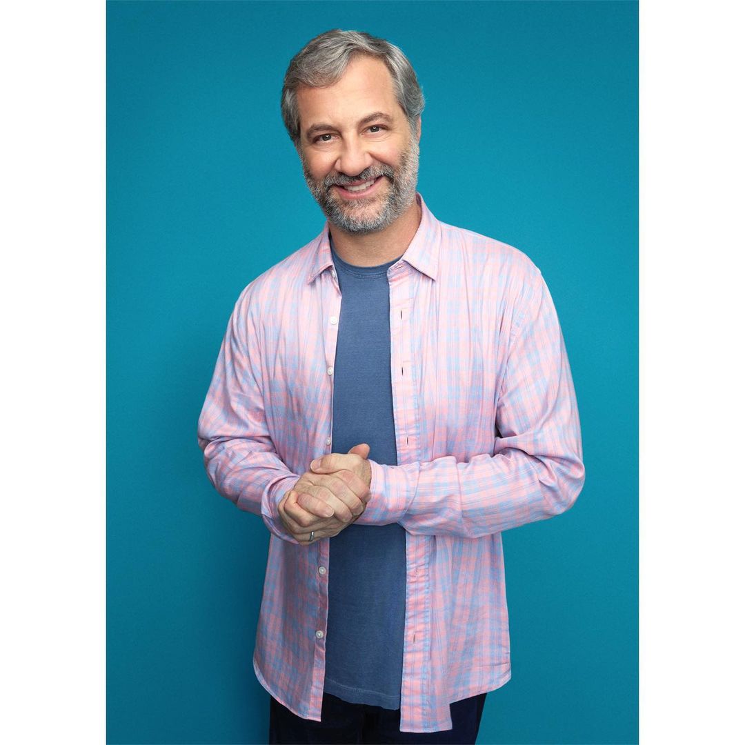 Portrait of smiling adult man wearing a pink and blue plaid shirt and a blue t-shirt standing against a teal background. 