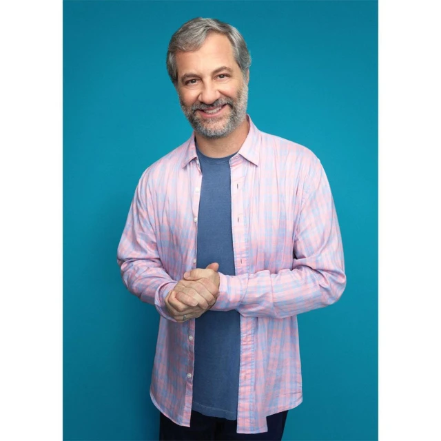 Portrait of smiling adult man wearing a pink and blue plaid shirt and a blue t-shirt standing against a teal background. 