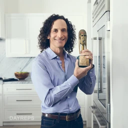 Man with long curly hair wearing a blue shirt holding a gold trophy inside a modern kitchen
