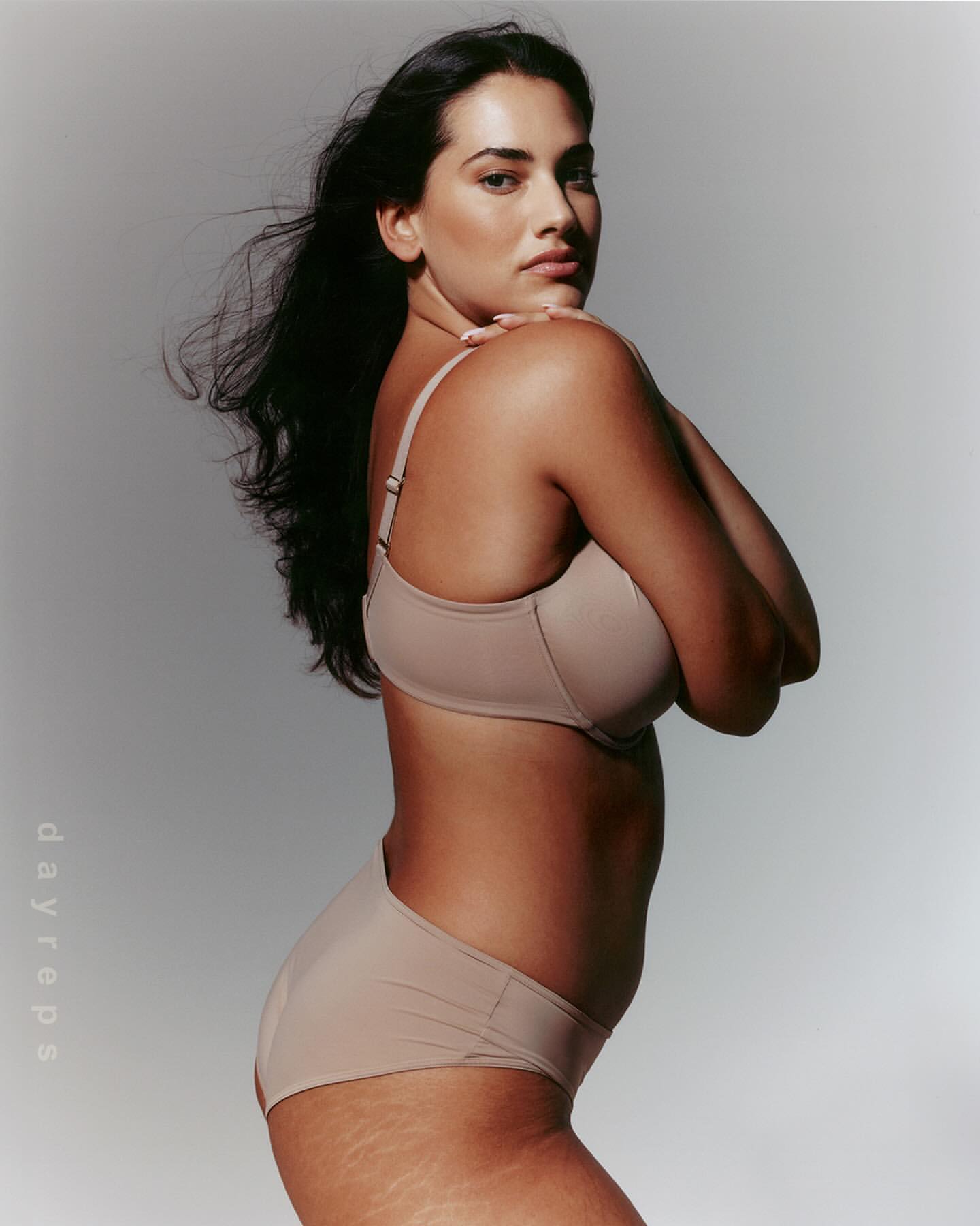 Woman poses in underwear and bra in a photo studio. The portrait has eye-level angle, neutral expression, and fair skin tone. The background is gray.