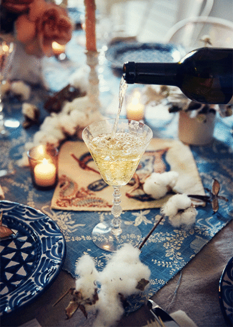 Pouring white wine into glass at table set with blue patterned cloth, plates, cotton sprigs, candles, flowers, and antique tablecloth.