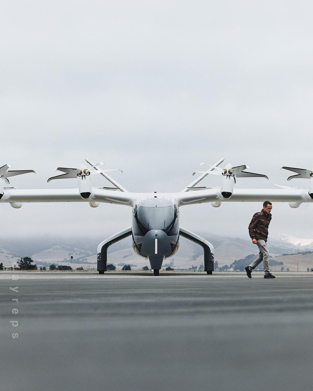 Silver eVTOL aircraft sits on the tarmac as a man walks by. A new form of personal air transportation on a cloudy day.