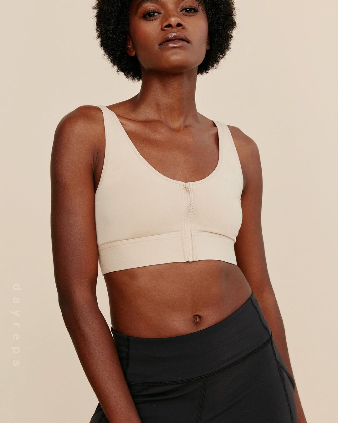 An adult female model poses in a photo studio wearing a beige sports bra and black leggings against a neutral-colored backdrop.