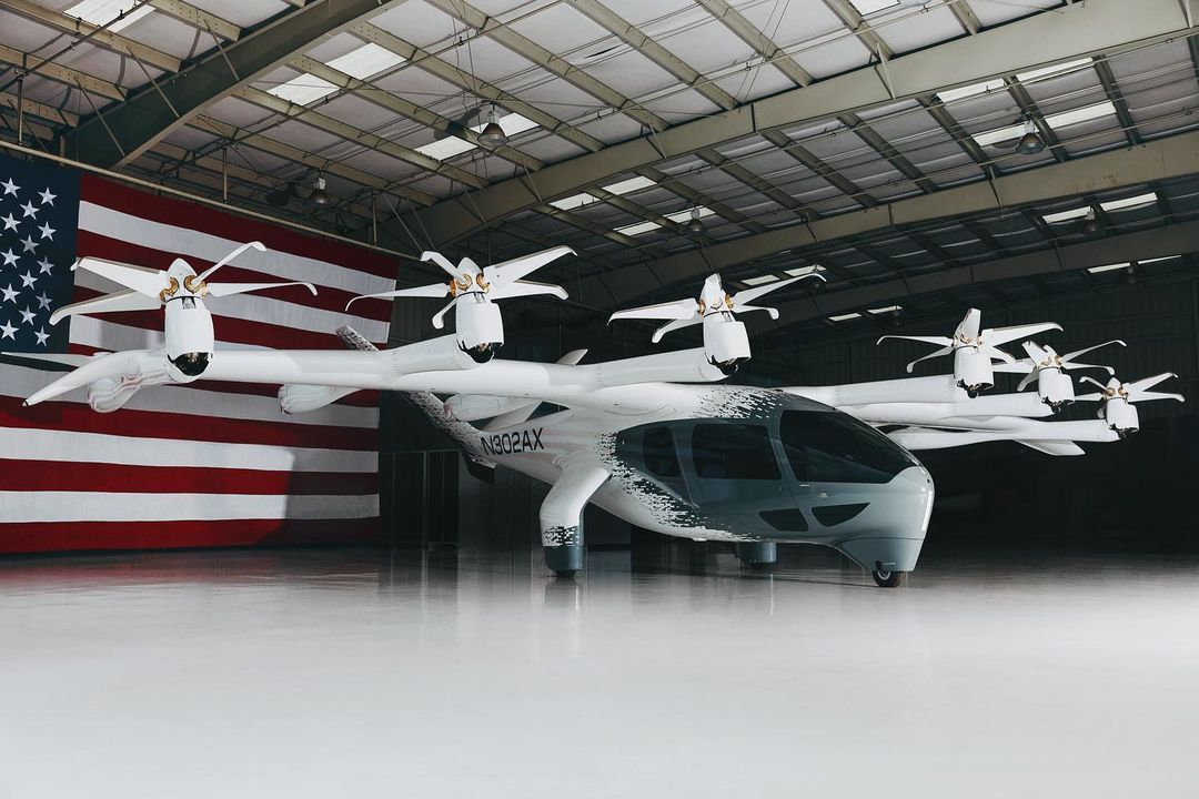White eVTOL aircraft with multiple rotors on display inside a hangar with an American flag in the background, well-lit indoor environment.