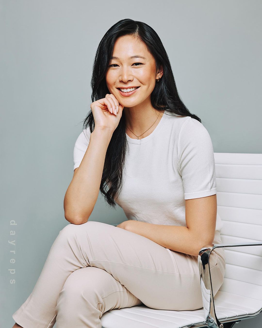 Smiling Asian woman sits on a modern white chair in a photo studio with a light gray background, wearing a white shirt and light pants.