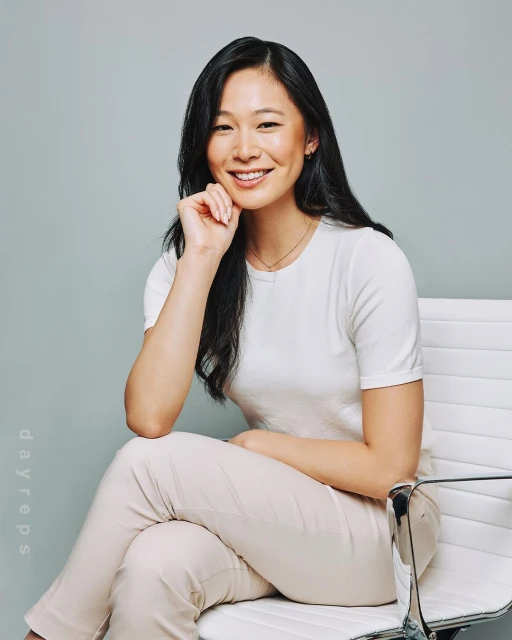 Smiling Asian woman sits on a modern white chair in a photo studio with a light gray background, wearing a white shirt and light pants.