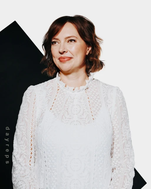 Portrait of an adult woman with short hair wearing a white lace top against a white backdrop with a black geometric shape.