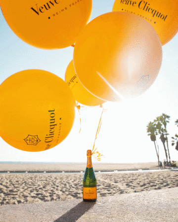 Celebratory scene on beach with Veuve Clicquot champagne bottle and bunch of branded yellow balloons against sky and palm trees.