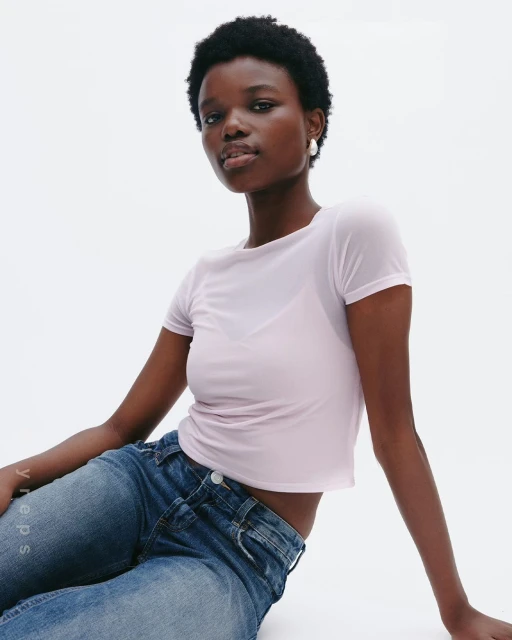 A Black woman sits in a photo studio wearing a light purple see-through shirt and blue jeans, looking directly at the camera.