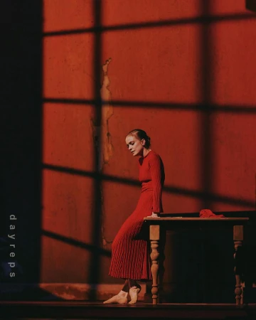 Woman wearing a red ribbed dress standing beside a wooden table against a dark red wall with distinct window shadow patterns