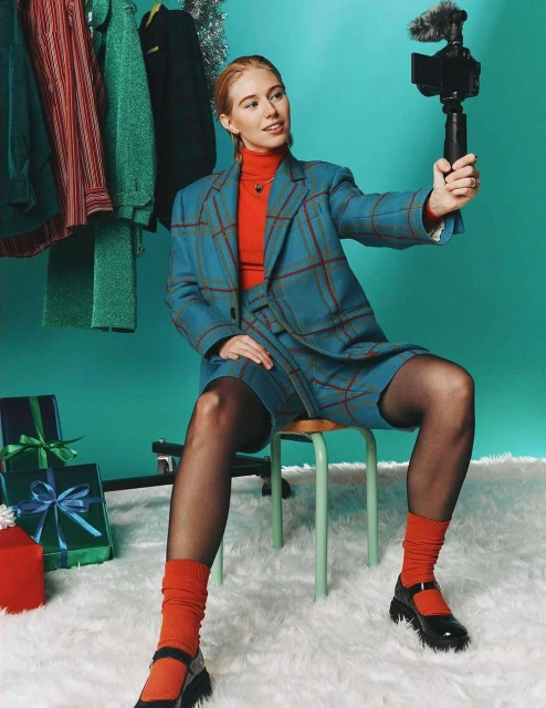 Young adult female vlogging with a camera sits on a stool against a teal background with gifts and clothing rack.