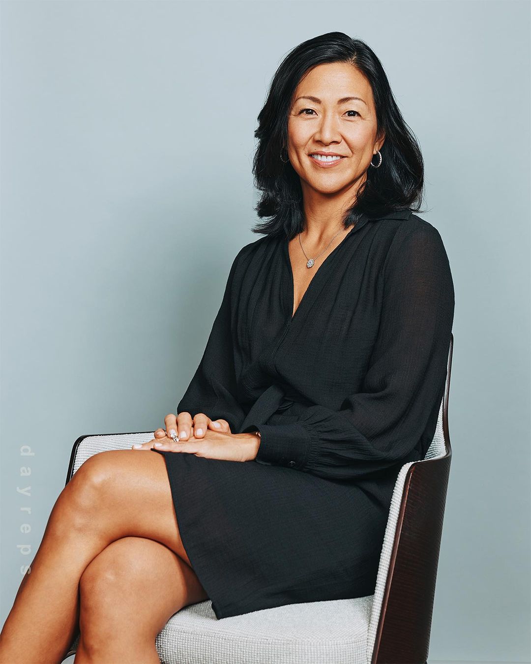 Portrait of a smiling, mature adult woman with dark hair, wearing a black dress and seated in a chair against a light-blue backdrop.