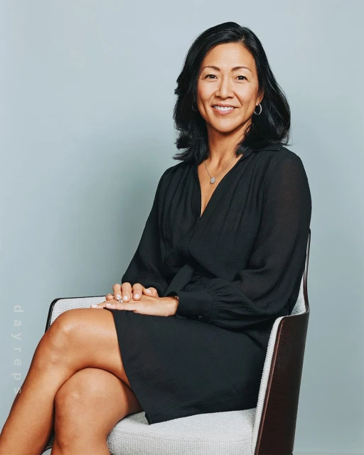 Portrait of a smiling, mature adult woman with dark hair, wearing a black dress and seated in a chair against a light-blue backdrop.