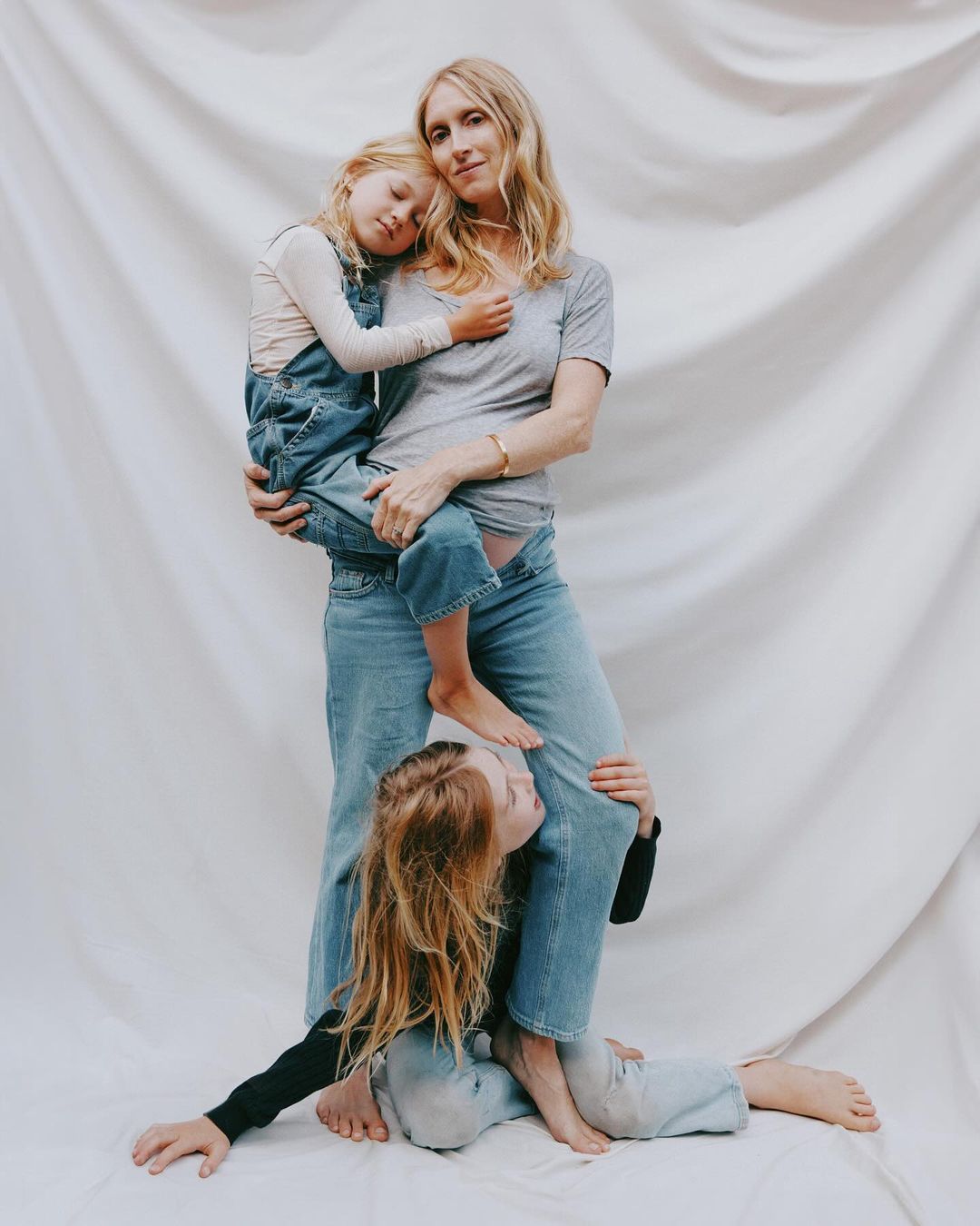 Woman holding a young child in denim overalls while another child sits at her feet against a draped white seamless backdrop.