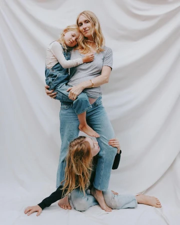 Woman holding a young child in denim overalls while another child sits at her feet against a draped white seamless backdrop.