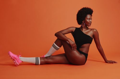 Athletic Black woman in a one-piece swimsuit and pink sneakers twists her body on an orange background. Fitness concept.