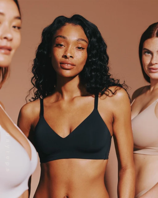 Three women of diverse ethnicities pose in bras against a neutral background in a close-up shot highlighting their natural beauty.