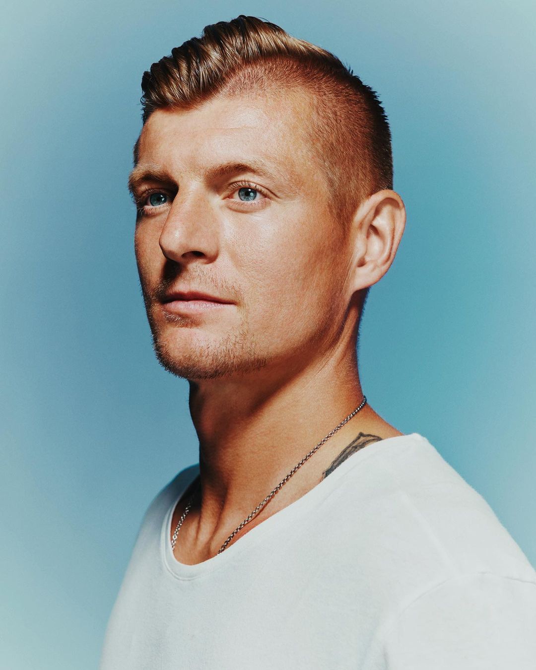 Portrait of an adult man with light brown hair, blue eyes, and a white t-shirt, looking off to the side against a light blue background.