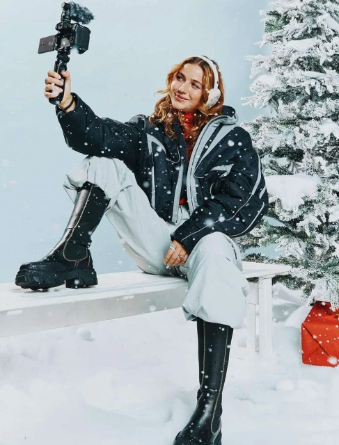 Young woman smiles while filming with camera. She sits on a snowy bench near a Christmas tree, wearing a black jacket and winter boots.
