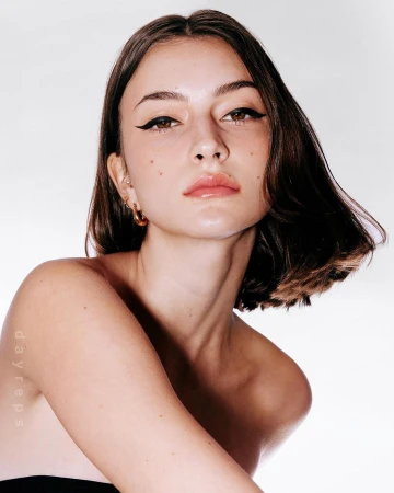 Close-up portrait of a woman with dark bobbed hair, winged eyeliner, and subtle freckles against a bright white background.