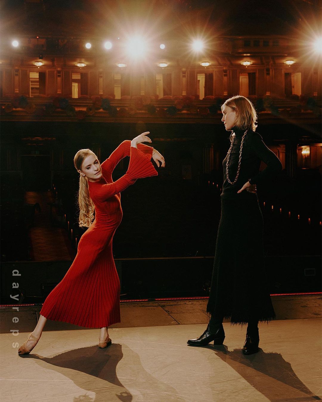 Two women on a stage. One woman in a red dress dances with arms outstretched, while the other in black stands and watches.
