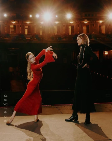 Two women on a stage. One woman in a red dress dances with arms outstretched, while the other in black stands and watches.