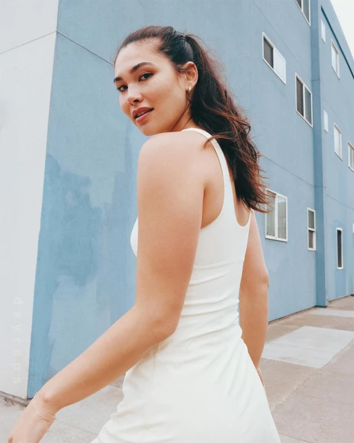 Adult female in a cream-colored dress poses outdoors with a light blue building in the background and looks over her shoulder.