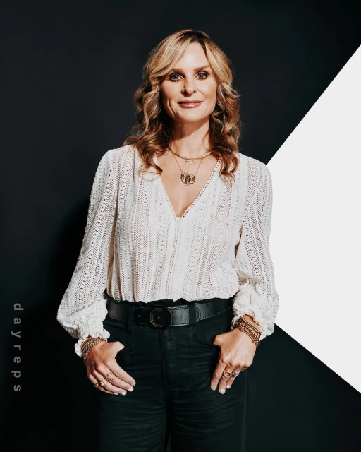 Adult woman with long blonde hair wearing a white lace blouse, black pants, belt and jewelry posing in front of a black and white wall.