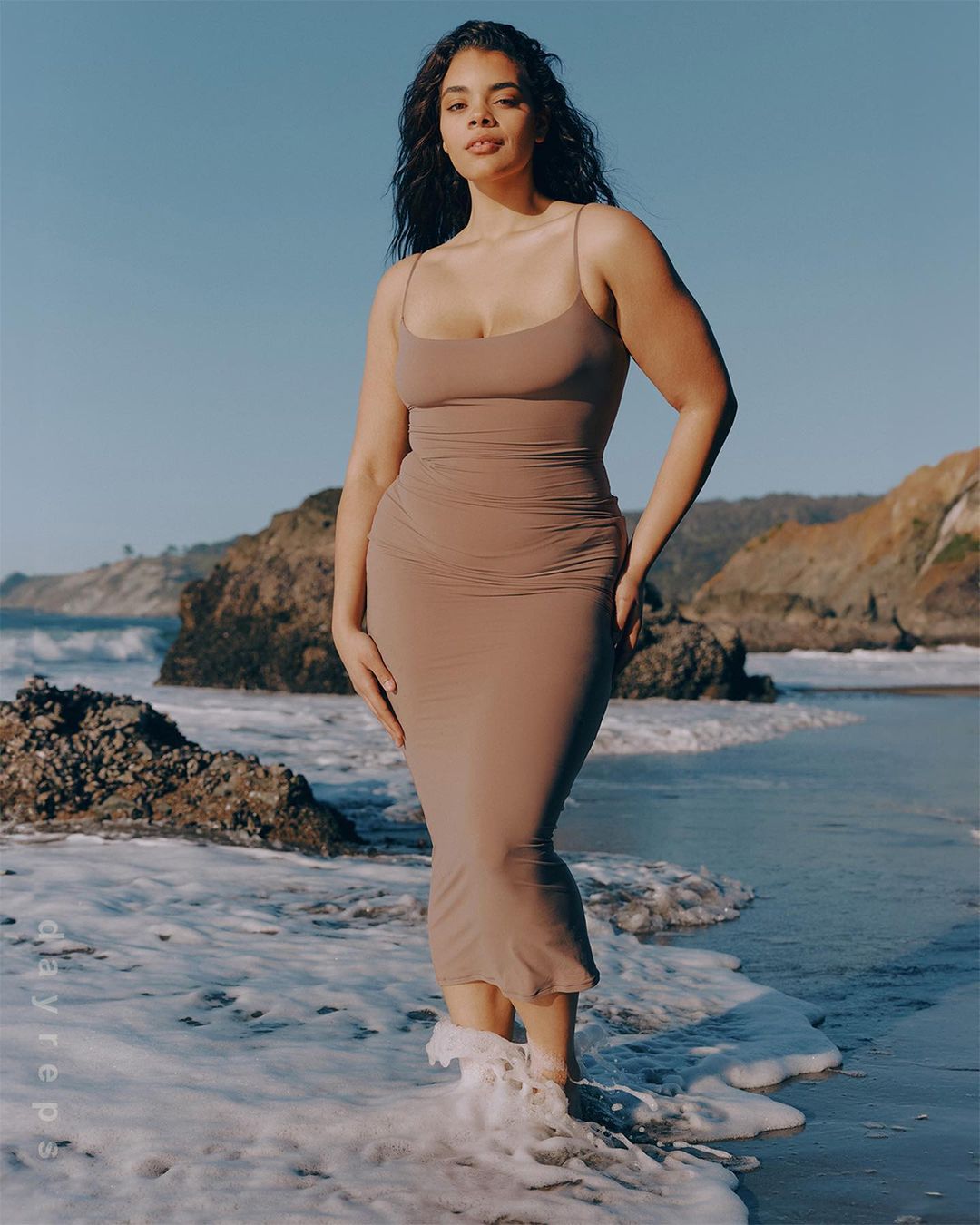 A tan-skinned woman in a beige dress stands in ocean waves on a beach with rocks and cliffs under a clear sky, looking at the camera.