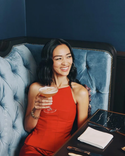 Brunette woman smiles while holding a cocktail. She wears a red dress and jewelry while sitting on a plush blue chair.