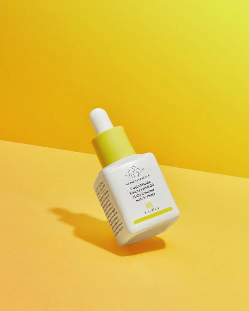 Drunk Elephant Virgin Marula Luxury Facial Oil in white bottle with yellow accents on a yellow background. Beauty and skincare product.