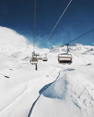 Ski lift chairs ascend over a snowy mountain landscape under a clear blue sky on a sunny day, providing transport for skiers.