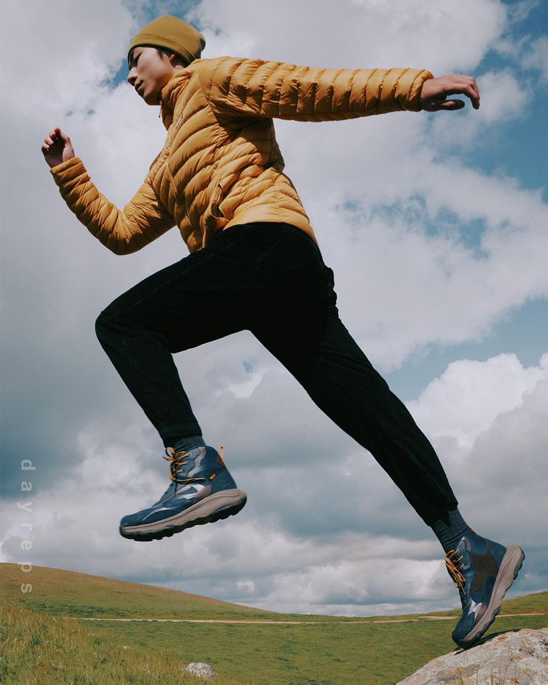 Man in a puffer jacket leaps from a rock in hiking boots against a cloudy sky and grassy hill, demonstrating outdoor activity.