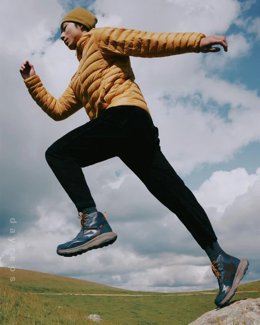 Man in a puffer jacket leaps from a rock in hiking boots against a cloudy sky and grassy hill, demonstrating outdoor activity.