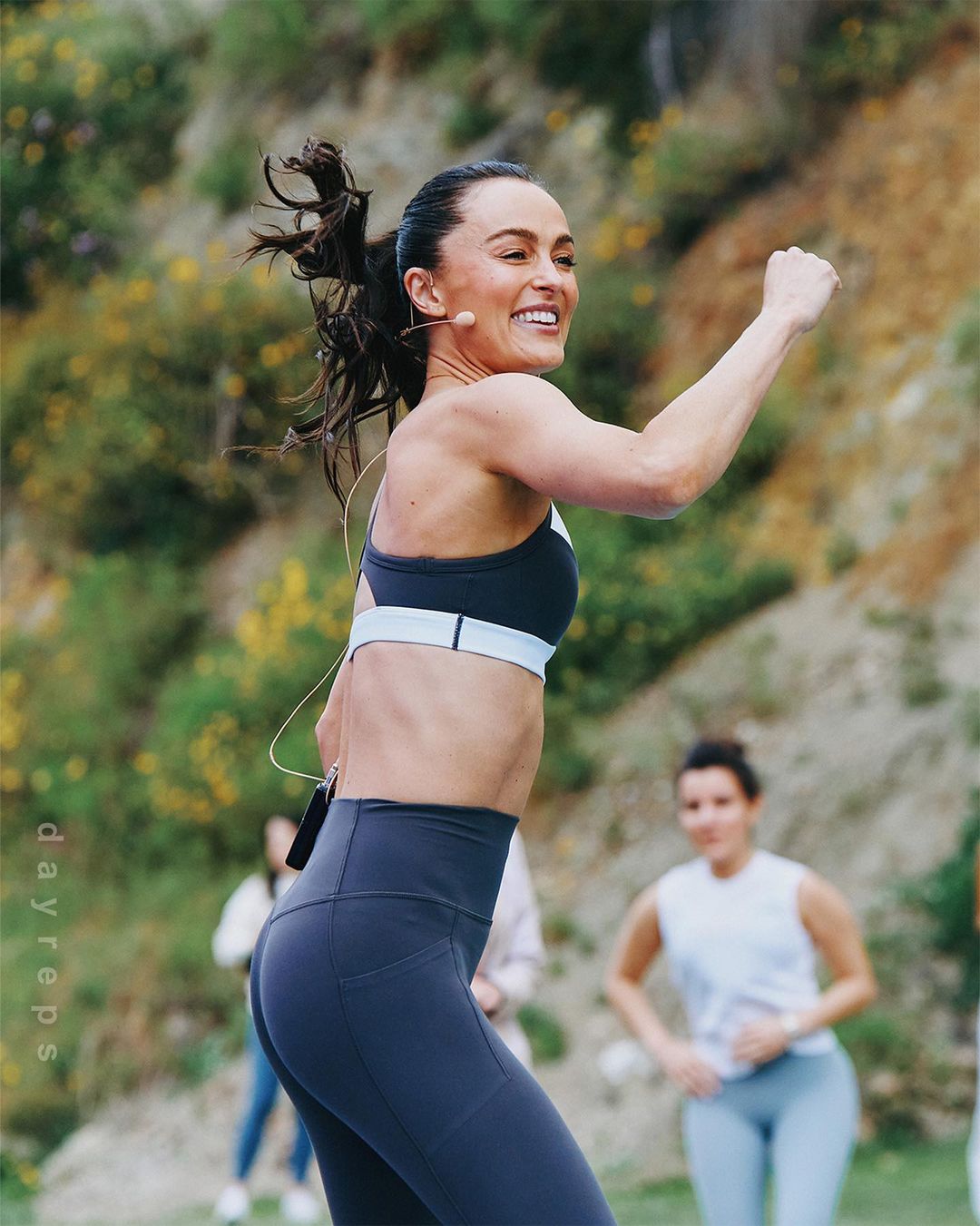Active woman in a sports bra and leggings smiles during outdoor exercise with a group in a grassy setting with a hillside backdrop.