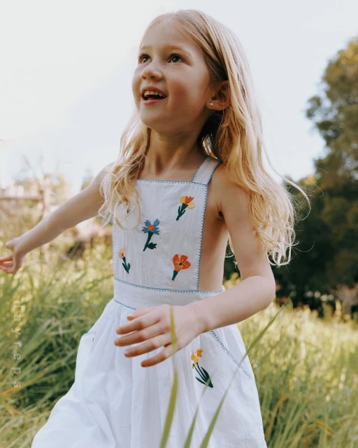 A young girl with blonde hair smiles while standing in a sunlit meadow, wearing a white dress adorned with embroidered flowers.