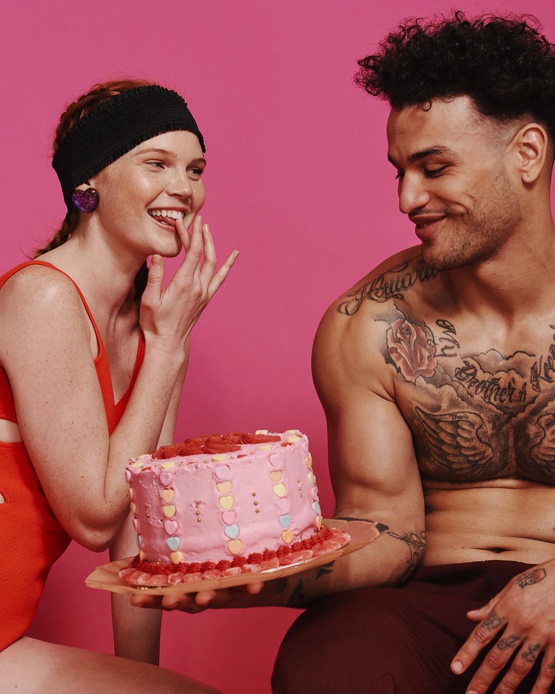 A couple shares a pink heart-decorated cake in a brightly lit studio portrait with a pink background, focusing on fashion and lifestyle.