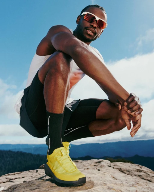 A man in athletic wear and sunglasses crouches on a rock showing off a yellow trail running shoe against a blue sky and mountain backdrop.