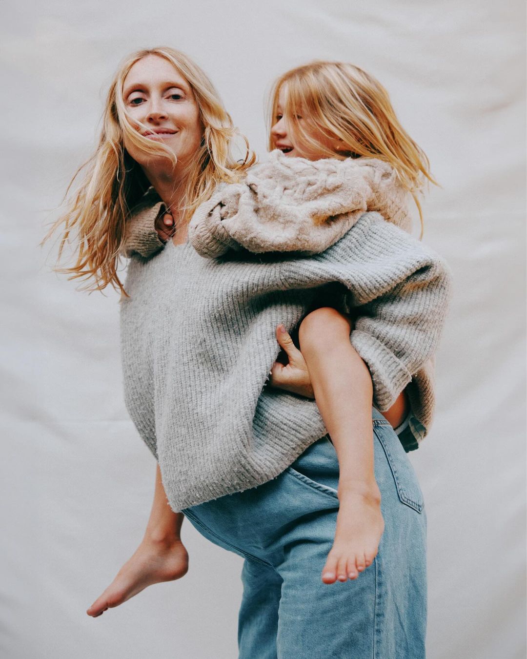 An adult female carries a child piggyback style. They are both fair-skinned with blond hair and wear casual clothing against a neutral backdrop.