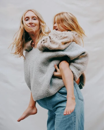 An adult female carries a child piggyback style. They are both fair-skinned with blond hair and wear casual clothing against a neutral backdrop.