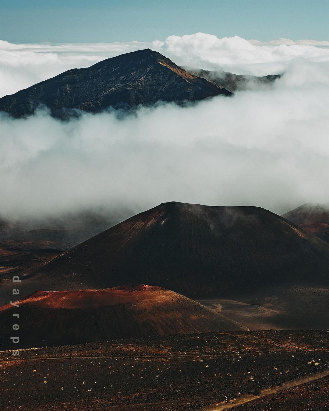 Dramatic aerial view of volcanic landscape with mountain peaks surrounded by thick clouds and desolate terrain in shades of brown and black.