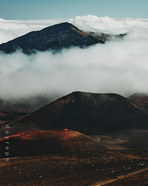 Dramatic aerial view of volcanic landscape with mountain peaks surrounded by thick clouds and desolate terrain in shades of brown and black.