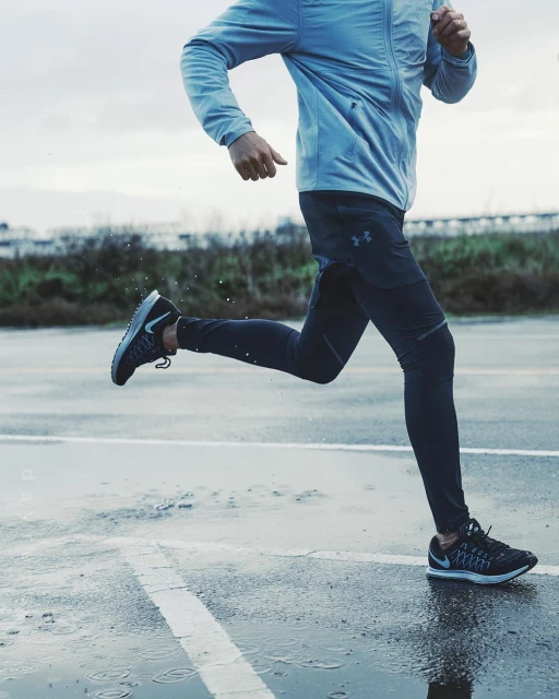 Man running on a wet paved surface. He is wearing sports clothing, including a jacket and running pants, against an overcast sky.