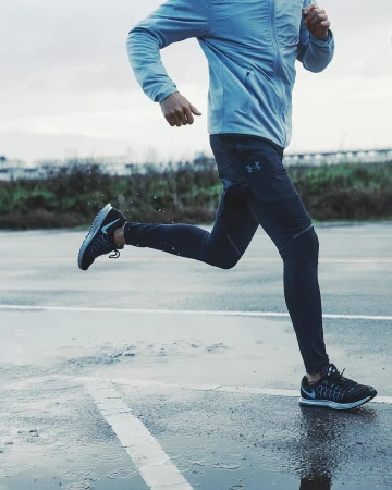 Man running on a wet paved surface. He is wearing sports clothing, including a jacket and running pants, against an overcast sky.