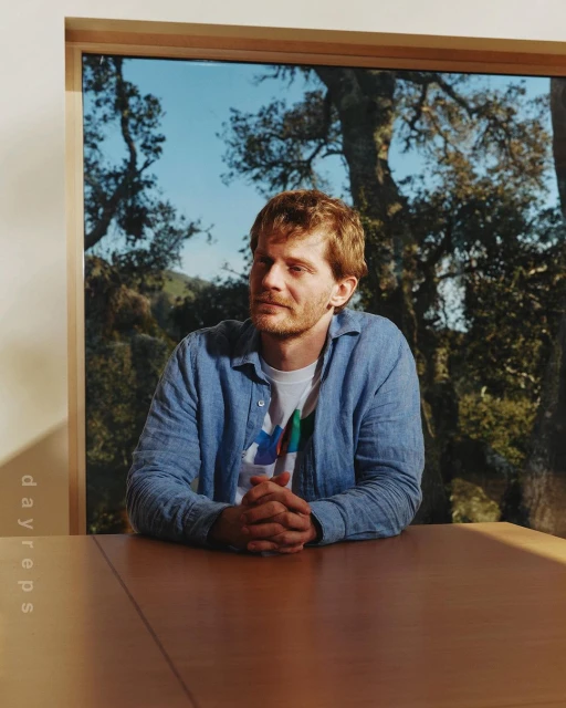 A man with light brown hair sits at a wooden table in front of a window with a forest scene on a bright day.