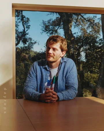 Man with reddish-brown hair wearing a blue button-down shirt seated at a wooden table with a window and trees behind him