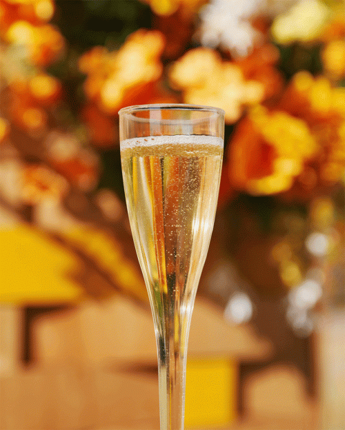 Close-up shot of a flute glass filled with a sparkling beverage, set against a blurred background of orange and white flowers.