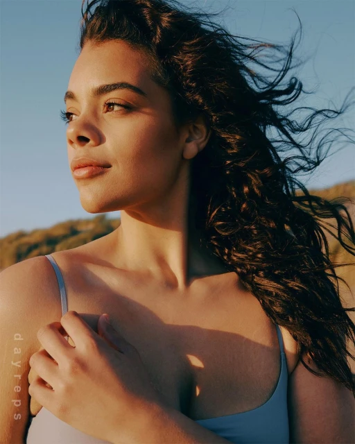 Portrait of a young woman with curly dark hair, wearing a light blue top, looking to the side, with a blurred background of land and sky.
