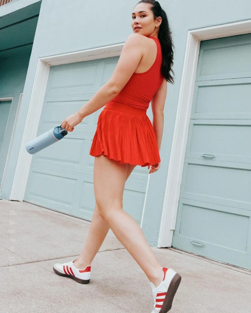 Athletic woman in red activewear walks outdoors holding a blue water bottle, wearing sneakers, against a light blue background.
