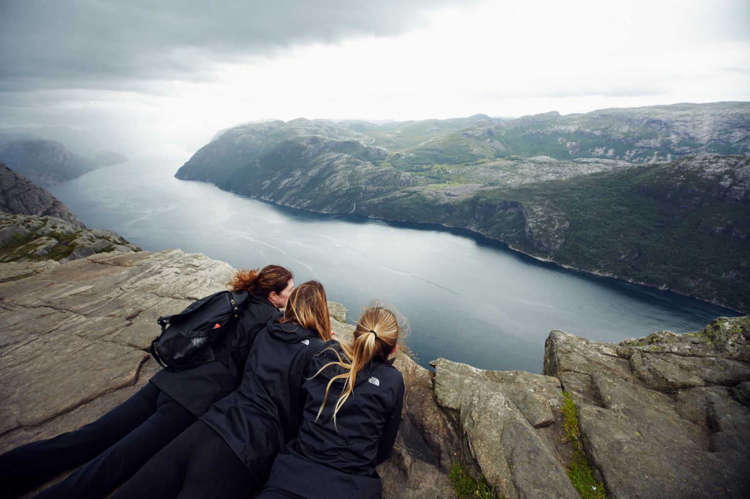 Three women overlook a fjord from a rocky cliff in Norway. The viewpoint features mountains, forest, and a body of water.